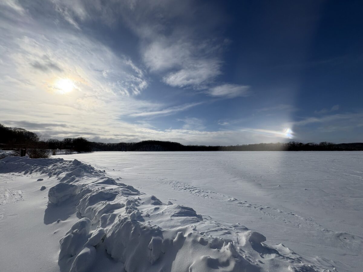 冬の網走。一面真っ白に凍りついた網走湖の雪原。太陽の光が氷晶に反射して現れる「幻日(サンドッグ)」が空に輝く幻想的な冬景色。
A breathtaking winter scenery of Lake Abashiri. A bright sun and a glowing parhelion (sun dog) shine over the pristine, untouched snow on the frozen lake under a blue sky.