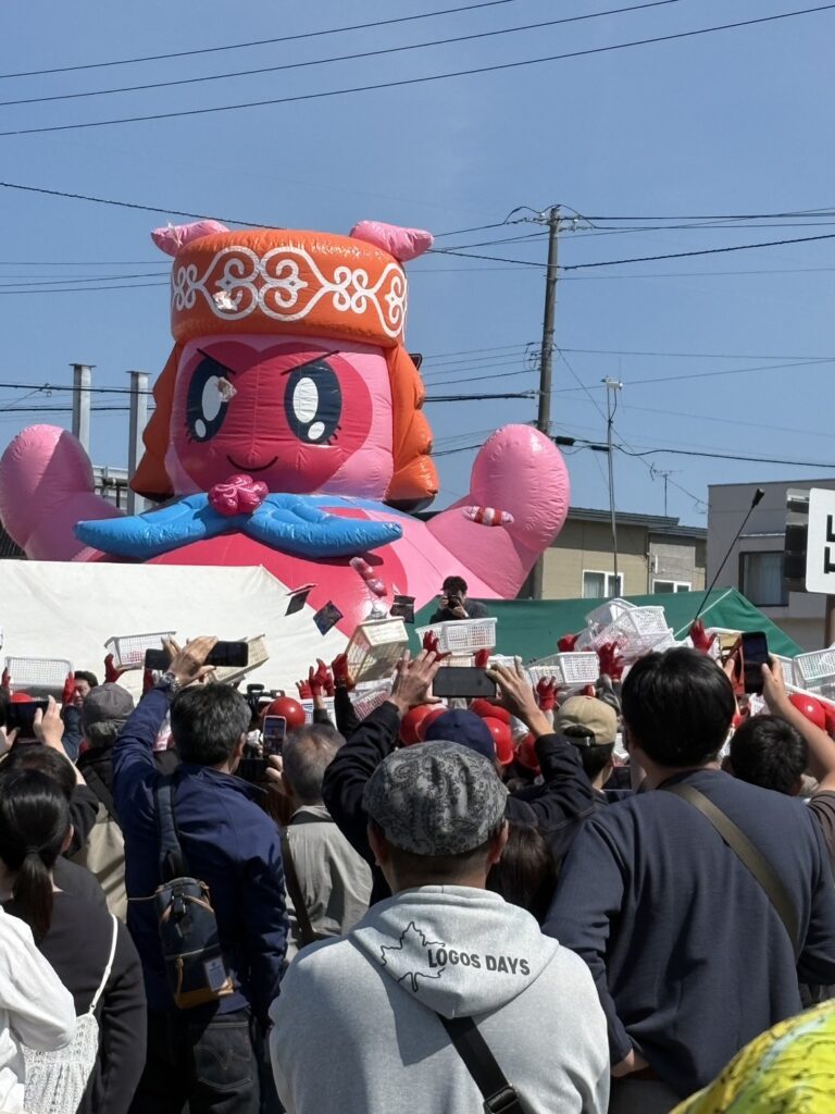 春カニ合戦in網走のメインイベントで、巨大なニポネのバルーンを背景に大勢の参加者が手を伸ばして盛り上がる会場の様子