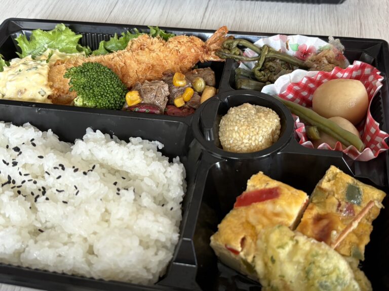 網走のデリカテッセン「ja.bitte（ビッテ）」の、大きなエビフライや肉料理、地元の旬の副菜がぎっしり詰まった豪華な特製お弁当 Top-down view of the luxurious and colorful "Tokusei Bento" featuring a large fried shrimp, meat, and seasonal side dishes at ja.bitte in Abashiri.