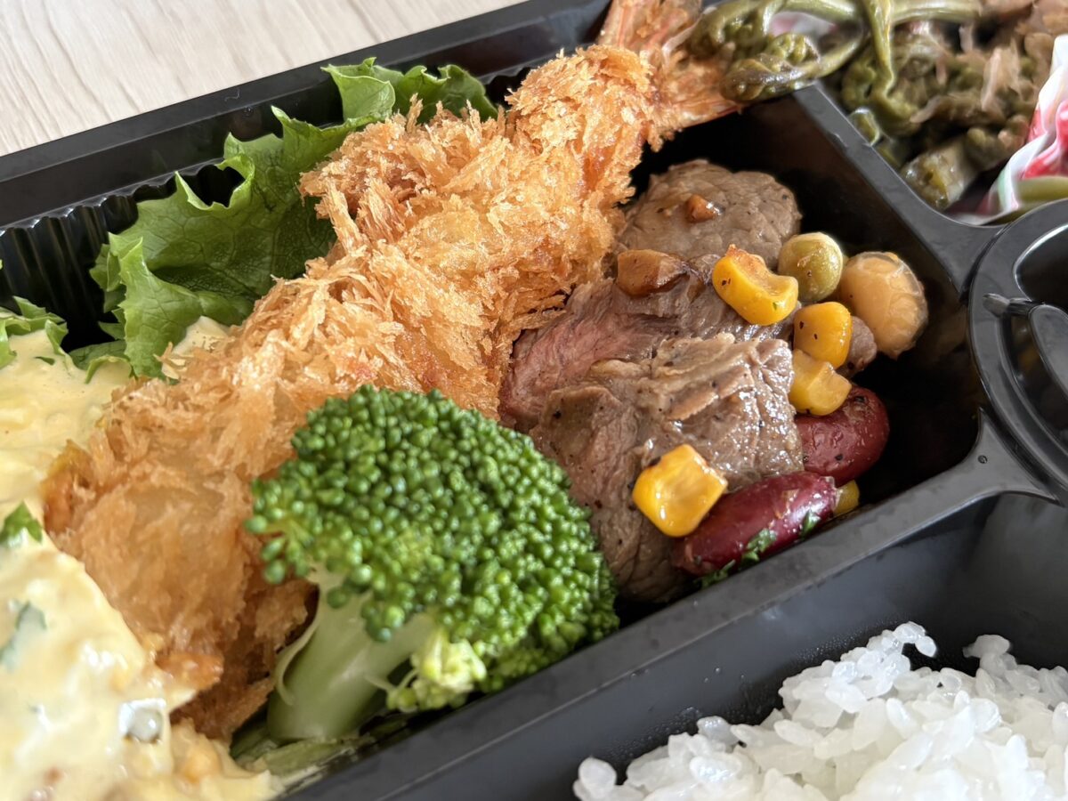 「ja.bitte（ビッテ）」のお弁当に入る、大きなサクサクのエビフライとジューシーなグリル肉のアップ
Close-up of a large crispy fried shrimp and juicy grilled meat in ja.bitte's bento box.