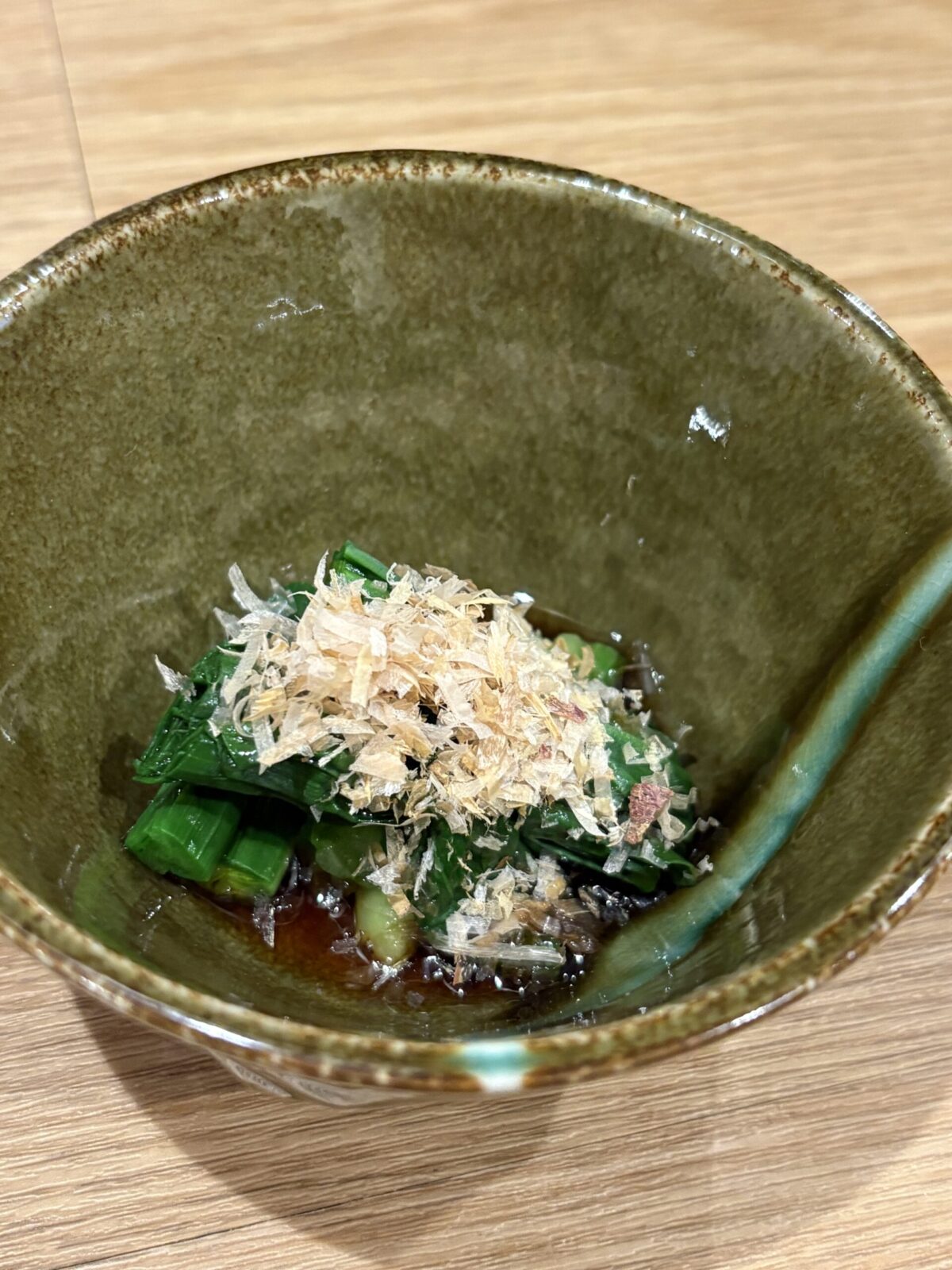 深緑色の和食器に大葉を敷き、鰹節（かつおぶし）を天盛りにした行者ニンニクのお浸し
Marinated wild alpine leeks (Gyoja-ninniku Ohitashi) placed on a shiso leaf and topped with bonito flakes in a ceramic bowl.