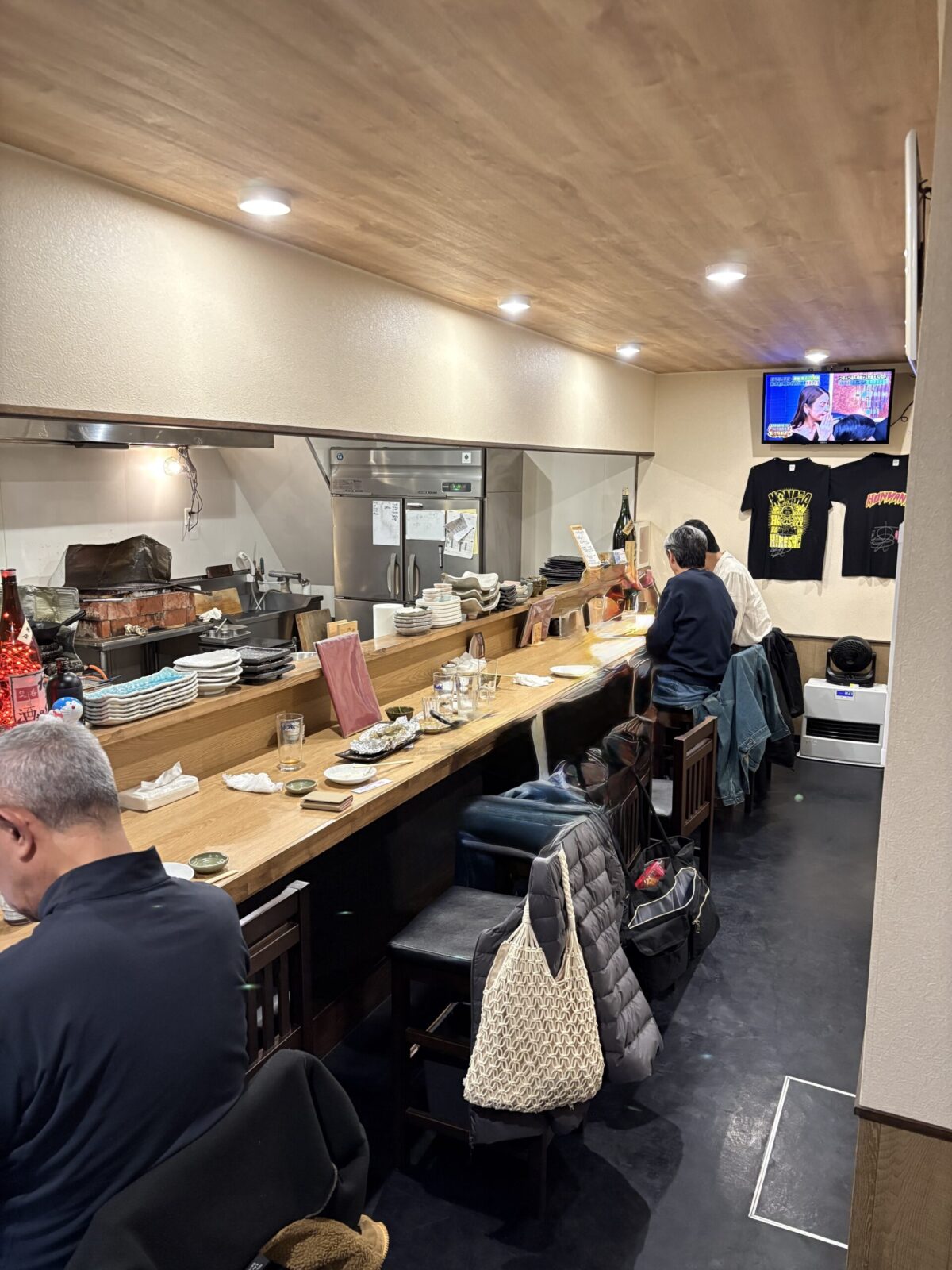網走「なかの酒場」の、厨房をぐるりと囲む一枚板の美しい木製カウンター席。常連客が寛ぎ、奥にはテレビや黒いオリジナルTシャツが飾られた温かみのある店内風景
The long wooden counter seating area inside Nakano Sakaba in Abashiri, creating a cozy and welcoming atmosphere for diners.