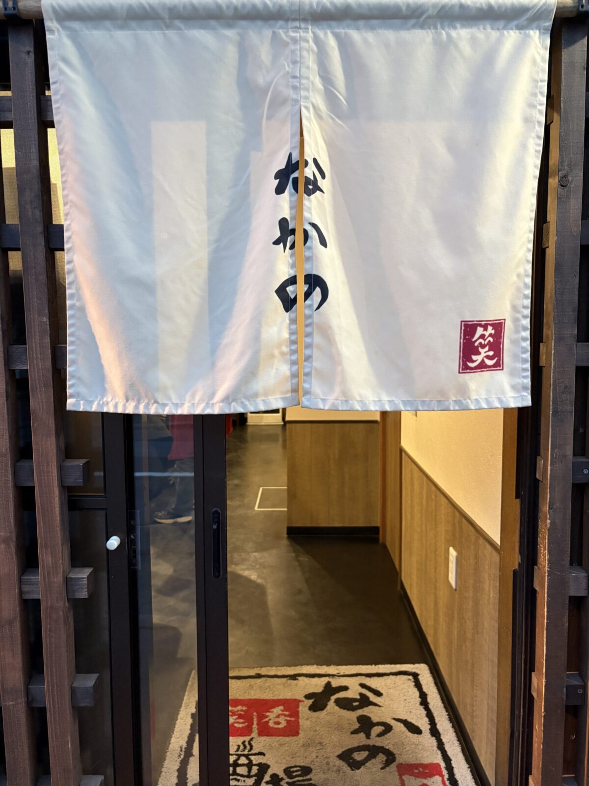 網走「なかの酒場」の入り口に掛かる、「なかの」の文字と赤い「笑」の刻印が入った白い暖簾（のれん）
Close-up of the white entrance curtain (noren) with the shop name at Nakano Sakaba in Abashiri.