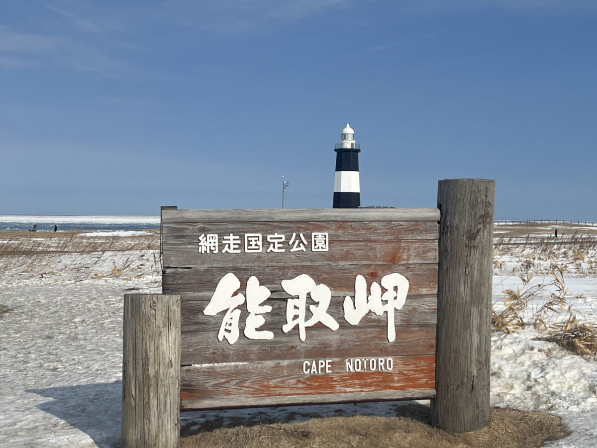 網走国定公園「能取岬」の味わい深い木製看板と、冬の雪景色に佇む白黒の灯台
The wooden sign of Abashiri Quasi-National Park "Cape Notoro" set against a snowy landscape with the lighthouse behind