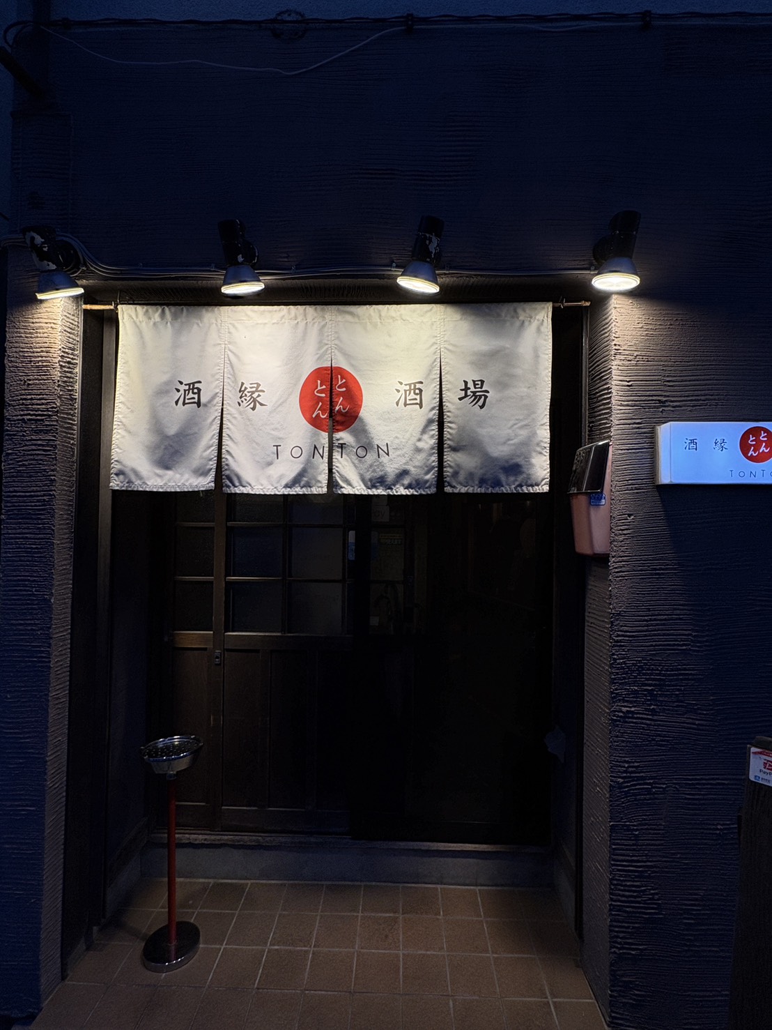 網走の居酒屋「酒縁酒場 屯屯(TONTON)」の夜の外観。白い暖簾とスポットライトに照らされた入口。 Night view of the entrance and noren curtain of izakaya "TONTON" in Abashiri, Hokkaido.